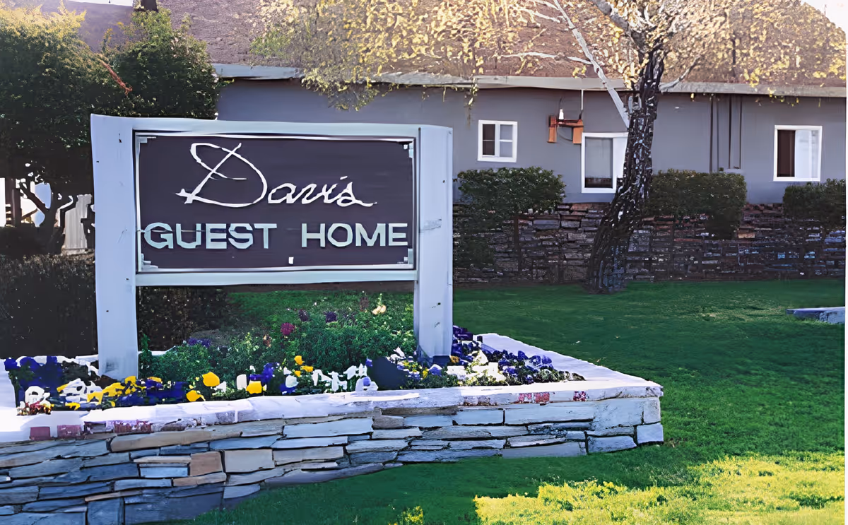 Outdoor view of a sign reading 'Davis Guest Home' in front of a building with a stone wall, green lawn, trees, and colorful flowers planted around the sign.