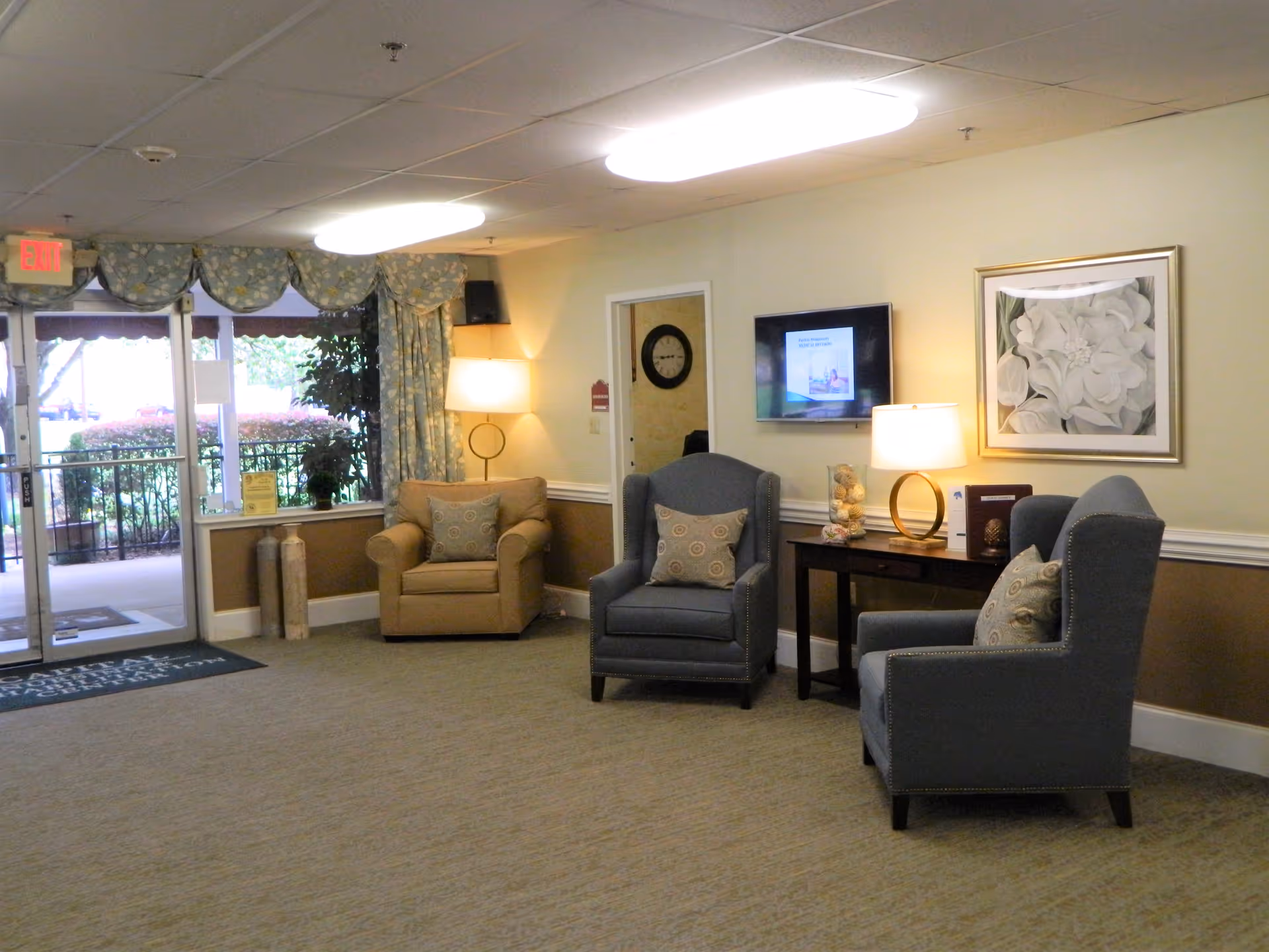 A cozy seating area in a nursing and rehabilitation center lobby with three upholstered armchairs arranged around a small table with a lamp and decorative items. A wall-mounted TV and framed floral artwork are visible on the wall. Large windows and glass doors let in natural light, and patterned curtains frame the windows.