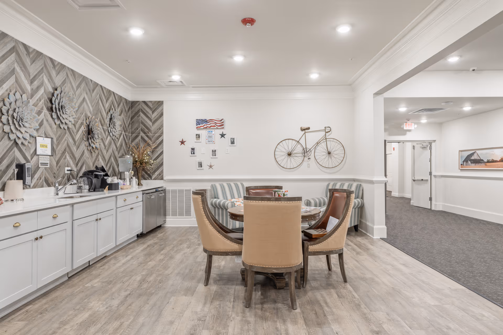 A bright and clean common area in Addison of Gwinnett Park featuring a round wooden table with four upholstered chairs, two striped armchairs against the wall, and a kitchenette with white cabinets, a sink, coffee maker, and water dispenser. The walls are decorated with a metal bicycle sculpture, an American flag, small framed pictures, and large flower wall art. The floor is light wood, and the ceiling has recessed lighting.