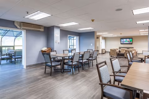 A spacious and well-lit common area in a senior living facility with multiple tables and chairs arranged for dining or socializing. The room has large windows allowing natural light, a television mounted on the far wall, and a doorway leading to an outdoor patio area with additional seating.