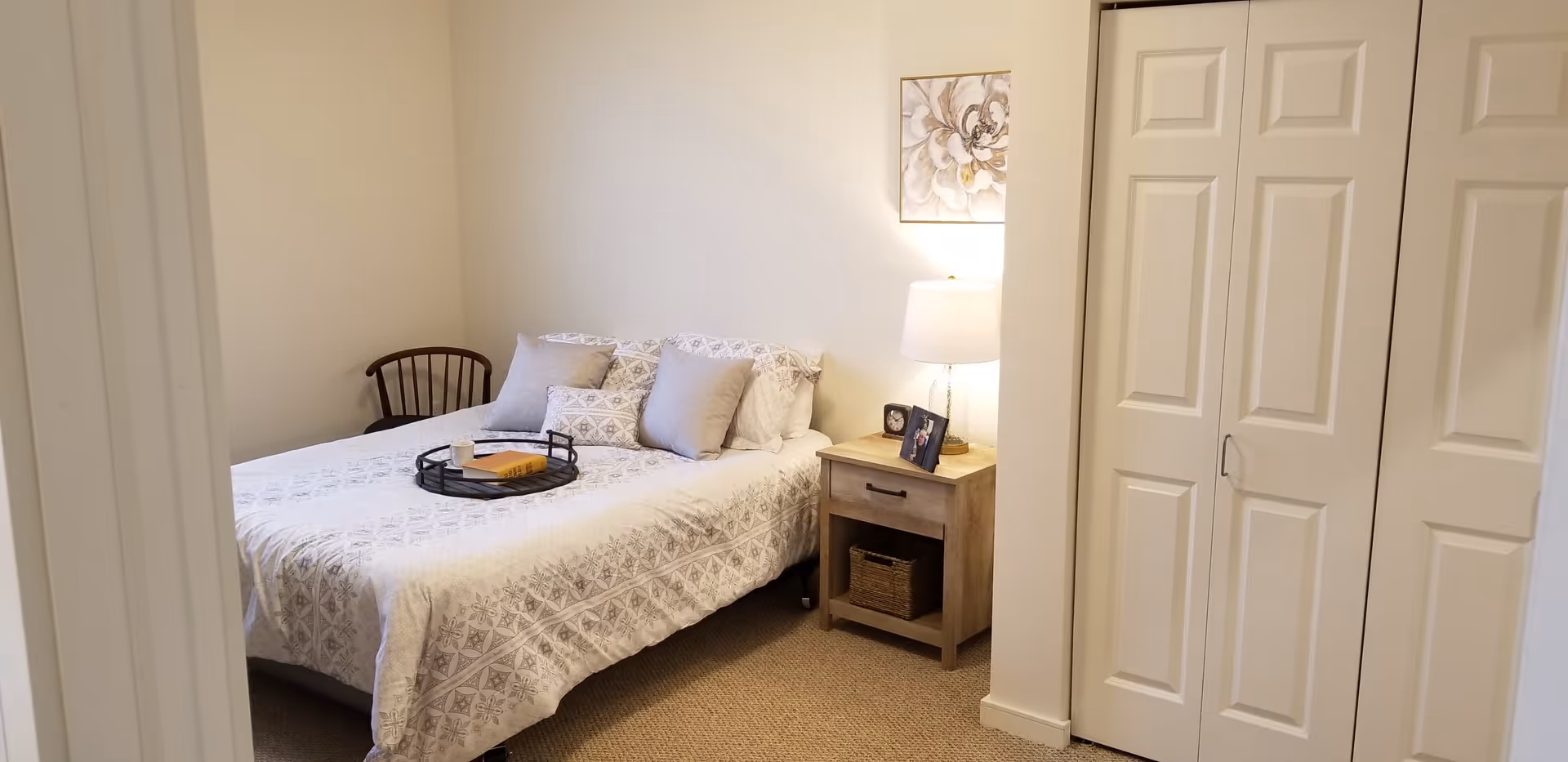 A cozy bedroom with a bed covered in a white patterned bedspread and several pillows. There is a wooden nightstand with a lamp, a framed photo, and a small clock. A wooden chair is placed in the corner, and a closet with white sliding doors is visible on the right side. A floral painting hangs on the wall above the nightstand.