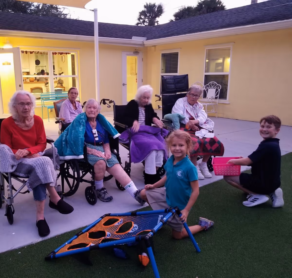 A group of elderly women, some in wheelchairs, sitting outside on a patio area of a yellow building during dusk. Two children, a girl kneeling and a boy sitting, are playing a bean bag toss game on the artificial grass in front of them. The scene appears to be a social or recreational activity at an assisted living facility.