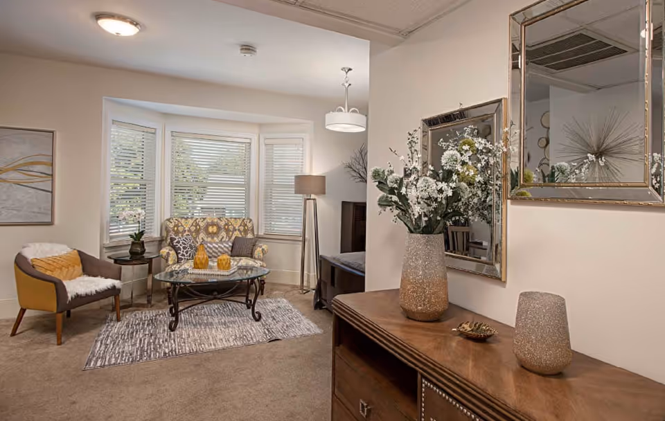 Cozy living room with a bay window, patterned loveseat and armchair around a glass coffee table, and a console table with decorative vase and mirror.