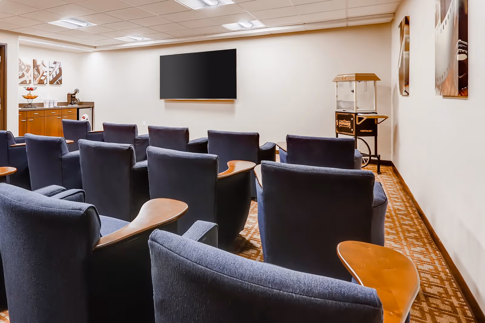 A small theater-style room with multiple rows of blue cushioned chairs equipped with wooden armrest trays, facing a large flat screen TV mounted on a white wall. To the left, there is a wooden cabinet with a coffee machine and cups, and to the right, a vintage popcorn machine on a stand. The room has a patterned carpet and three abstract art pieces hanging on the walls.