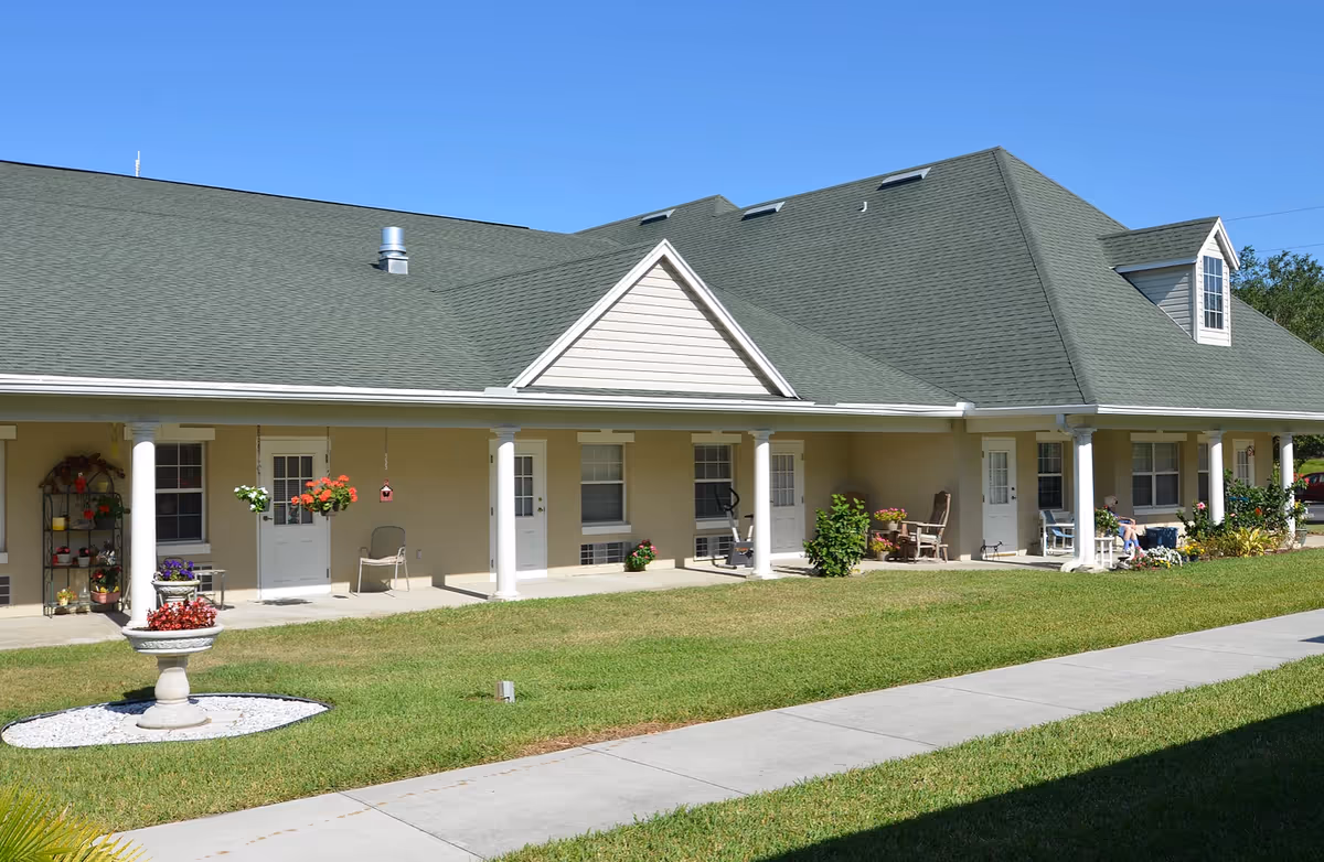 Exterior view of a single-story building with a green roof and beige walls, featuring a covered porch supported by white columns. The porch has several doors and windows, with chairs and potted plants placed along it. A well-maintained lawn and a concrete walkway are visible in the foreground under a clear blue sky.