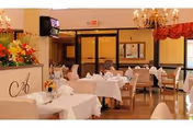 A dining room with tables covered in white tablecloths, set with napkins and glassware. There are wooden chairs around the tables, floral arrangements on a counter, chandeliers hanging from the ceiling, and large windows with red valances.