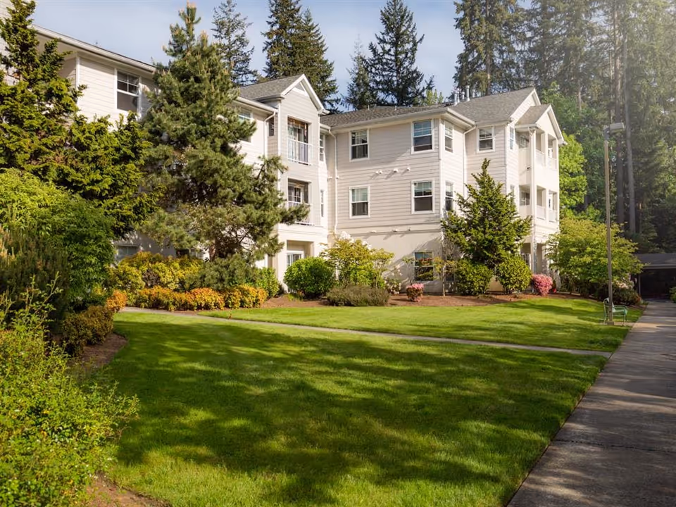 Exterior view of a multi-story senior living facility building surrounded by well-maintained green lawns, shrubs, and tall trees under a clear sky.