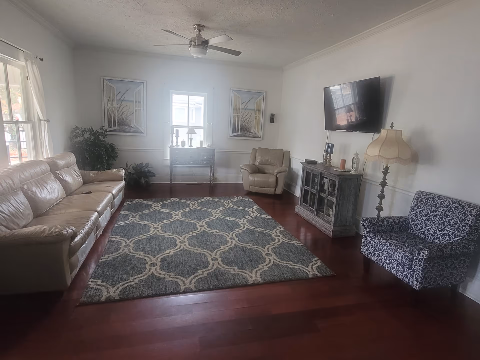 A living room with a beige leather sofa, a beige armchair, a patterned blue armchair, a large patterned rug on wooden floor, a wall-mounted flat screen TV above a rustic cabinet, a standing lamp with a beige shade, two framed pictures on the wall, a small table with decorative items in front of a window, and a ceiling fan.