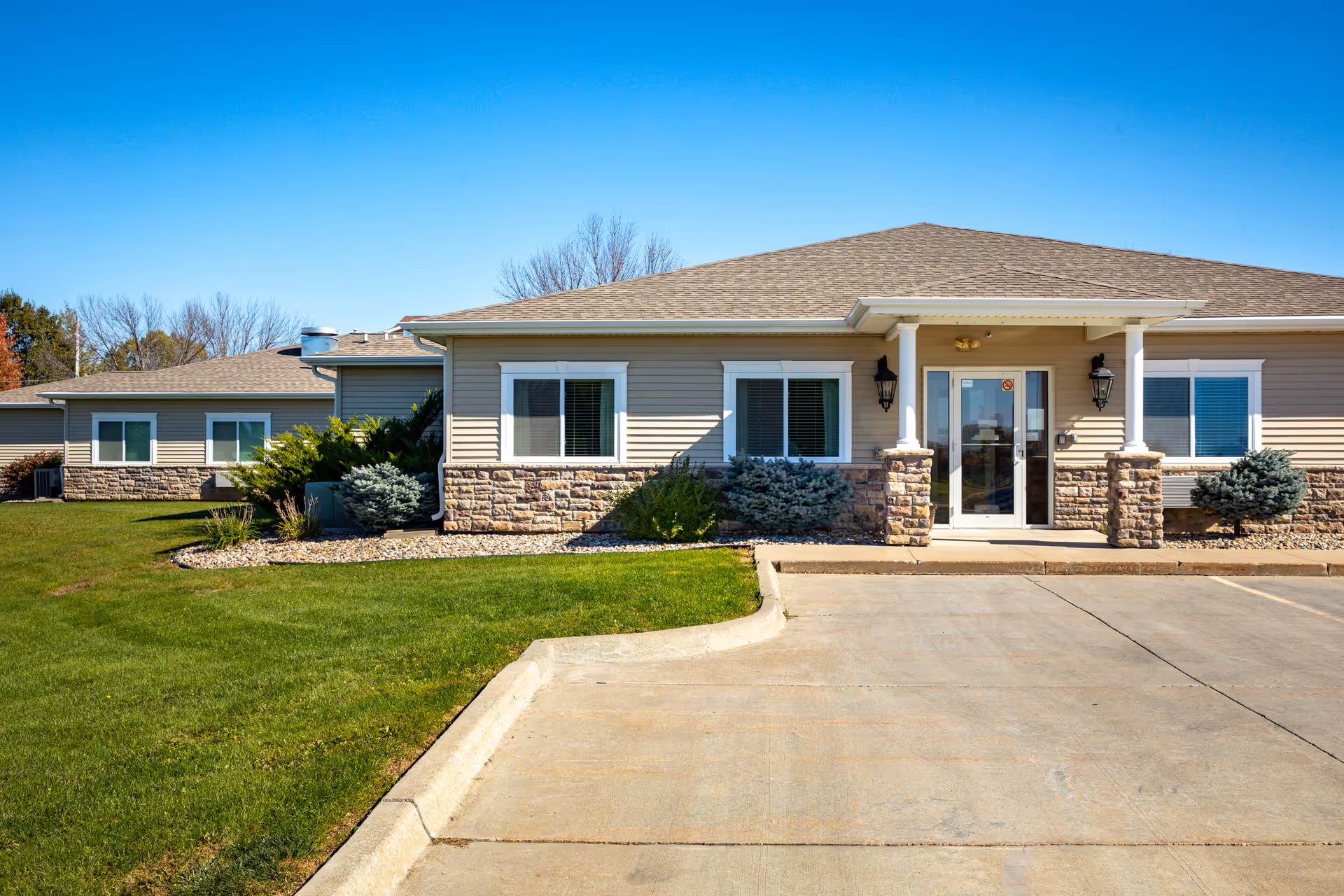 Exterior view of a single-story building with beige siding and stone accents, featuring a covered entrance with two columns and glass double doors. The building is surrounded by green grass, shrubs, and a clear blue sky.