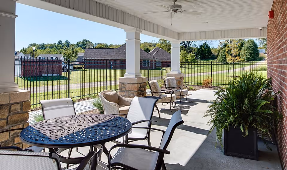 Covered outdoor patio area with a round metal table and several chairs. There are additional cushioned chairs along the side near a brick wall and large potted plants. The patio overlooks a fenced grassy area with trees and buildings in the background under a clear blue sky.