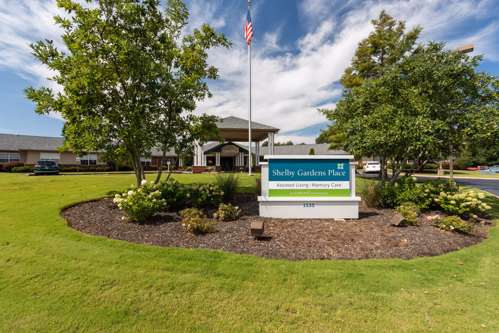 Front exterior view of Shelby Gardens Place assisted living and memory care facility with a landscaped garden, trees, an American flag on a flagpole, and a sign displaying the facility name and address.