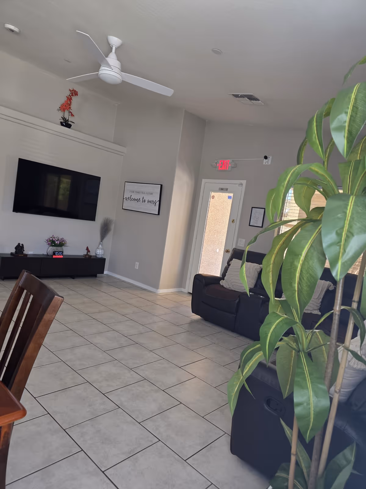 Bright common living room with tiled floor, wall-mounted TV, recliner sofas, and a large indoor plant near an exit door.