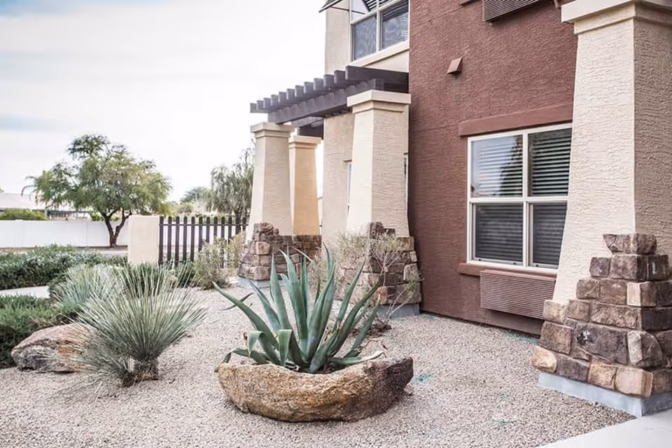 Exterior view of a building with beige and brown walls, stone pillars, and a small landscaped area with desert plants and gravel in front.