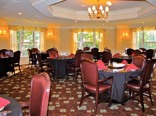 A dining room with round tables covered in black tablecloths, each set with red folded napkins, plates, and silverware. The room has large windows with yellow curtains, a patterned carpet, and a chandelier hanging from a tray ceiling.