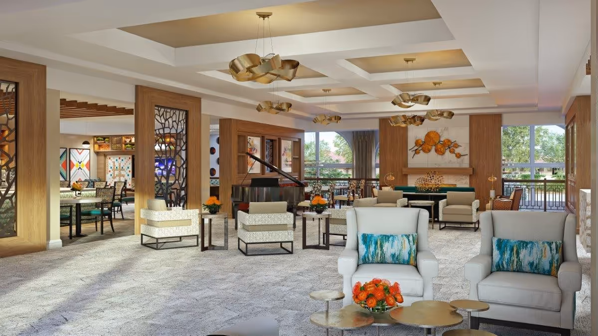A spacious and well-lit senior living facility common area with comfortable armchairs, small tables with orange flower arrangements, a grand piano, and decorative wooden partitions. The room features large windows allowing natural light and views of greenery outside, modern ceiling lights, and colorful artwork on the walls.