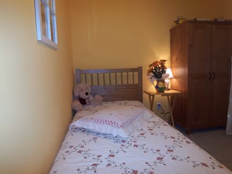 A small bedroom with yellow walls featuring a single bed with floral bedding and a pillow. A teddy bear is placed at the head of the bed. Next to the bed is a wooden side table with a lamp and a vase of flowers. A wooden wardrobe is positioned against the wall in the corner of the room.