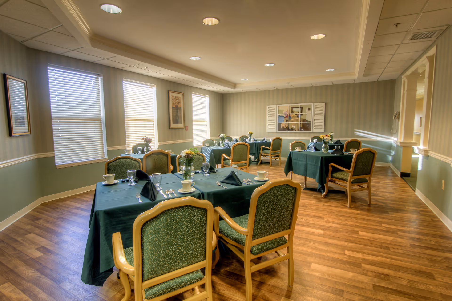 A dining room in a senior living facility with several tables covered in green tablecloths, each set with cups, glasses, silverware, and napkins. The room has wooden flooring, green and beige walls, three large windows with blinds, framed artwork on the walls, and ceiling lights.