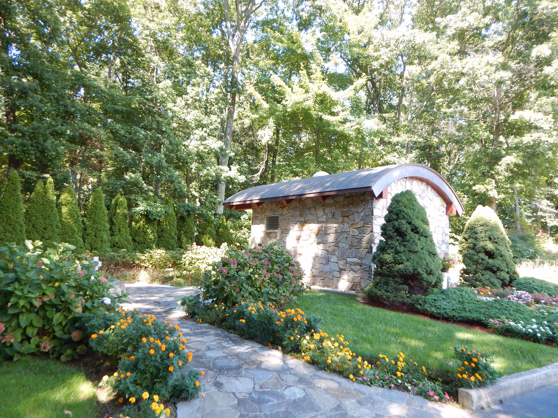 A stone pathway winds through a well-maintained garden with vibrant orange and pink flowers, green shrubs, and tall trees. A small stone building with a curved roof is visible in the background, surrounded by lush greenery.