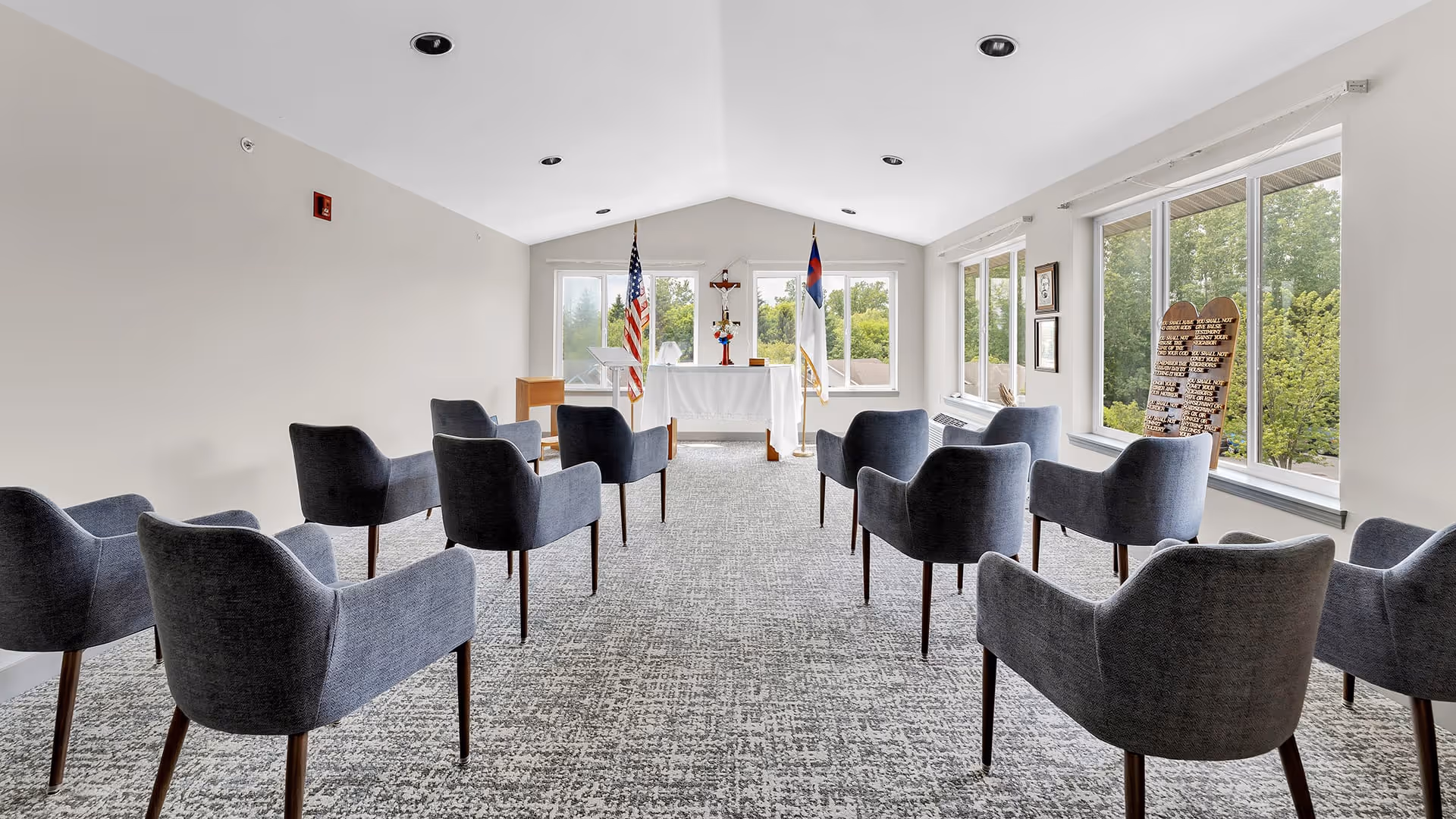 A small chapel or prayer room with gray upholstered chairs arranged in rows facing a white altar. Behind the altar are two flags, one American and one Christian, and a crucifix mounted on the wall. Large windows on the right side let in natural light and show green trees outside. The room has a light gray carpet and white walls.