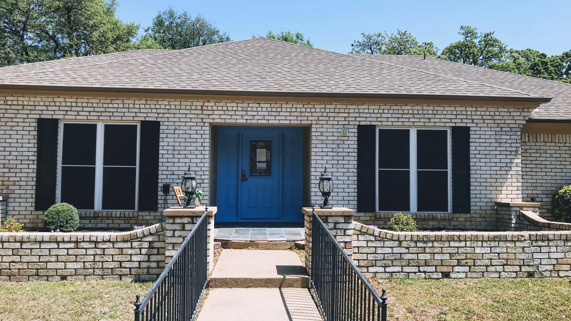 Front exterior view of a single-story brick building with a blue door, black window shutters, and a small garden area bordered by a low brick wall and black metal railings leading up to the entrance.