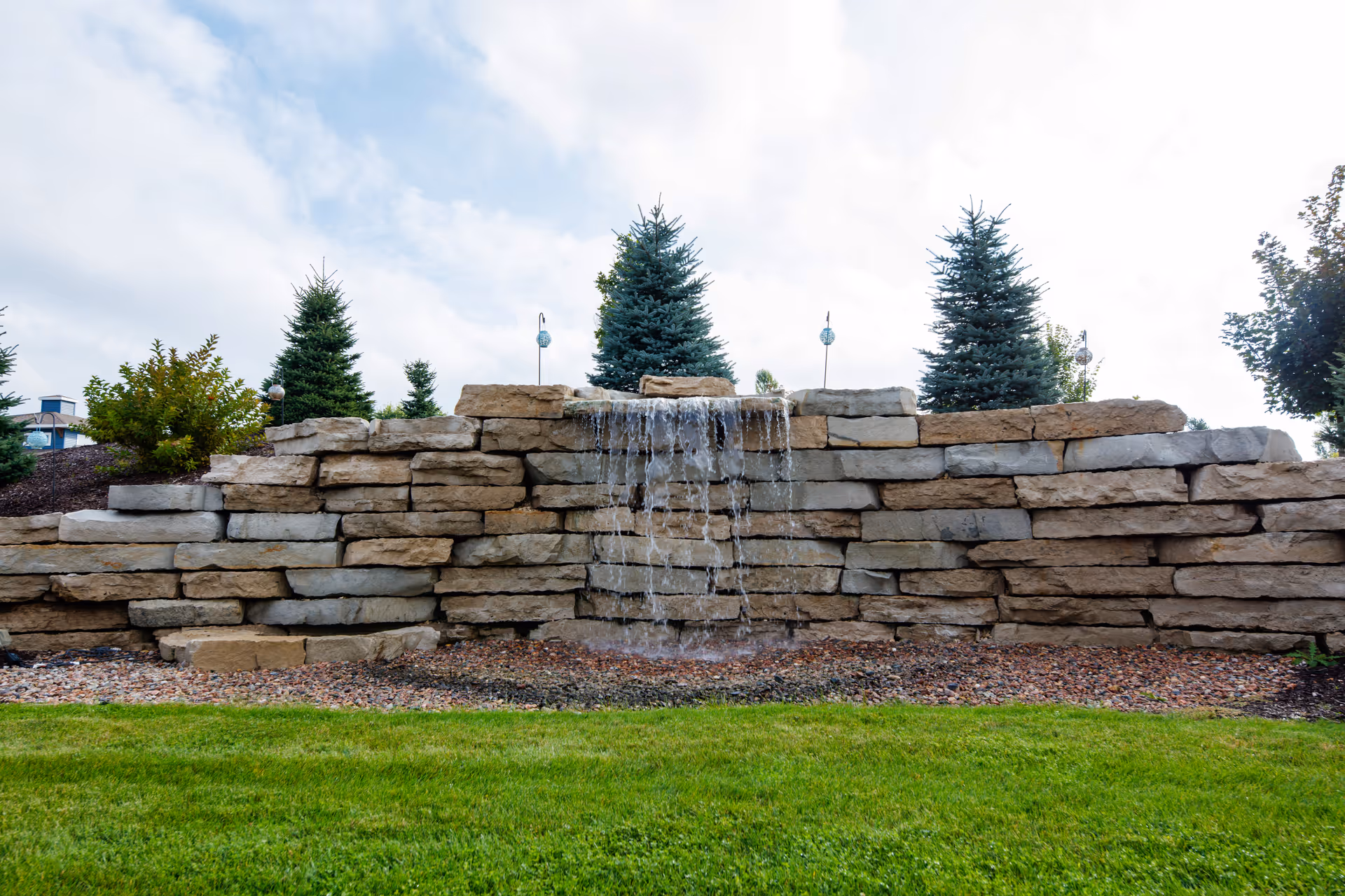 Stone retaining wall with a small waterfall feature, evergreen trees behind it, and a grass lawn in front.