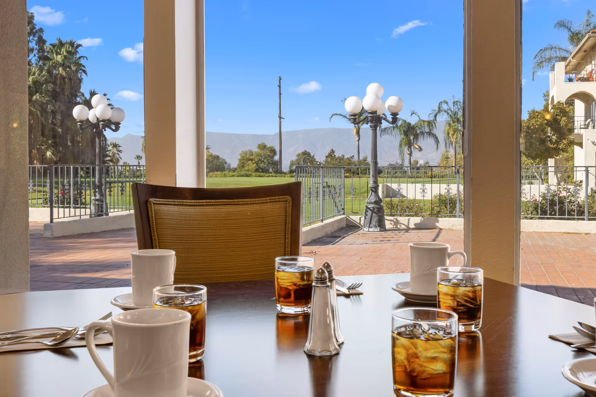 A dining table set with four glasses of iced tea, four white coffee mugs, silverware, and a salt shaker. The table is indoors near large windows that offer a view of an outdoor patio with lamp posts, palm trees, green grass, and distant mountains under a blue sky with a few clouds.