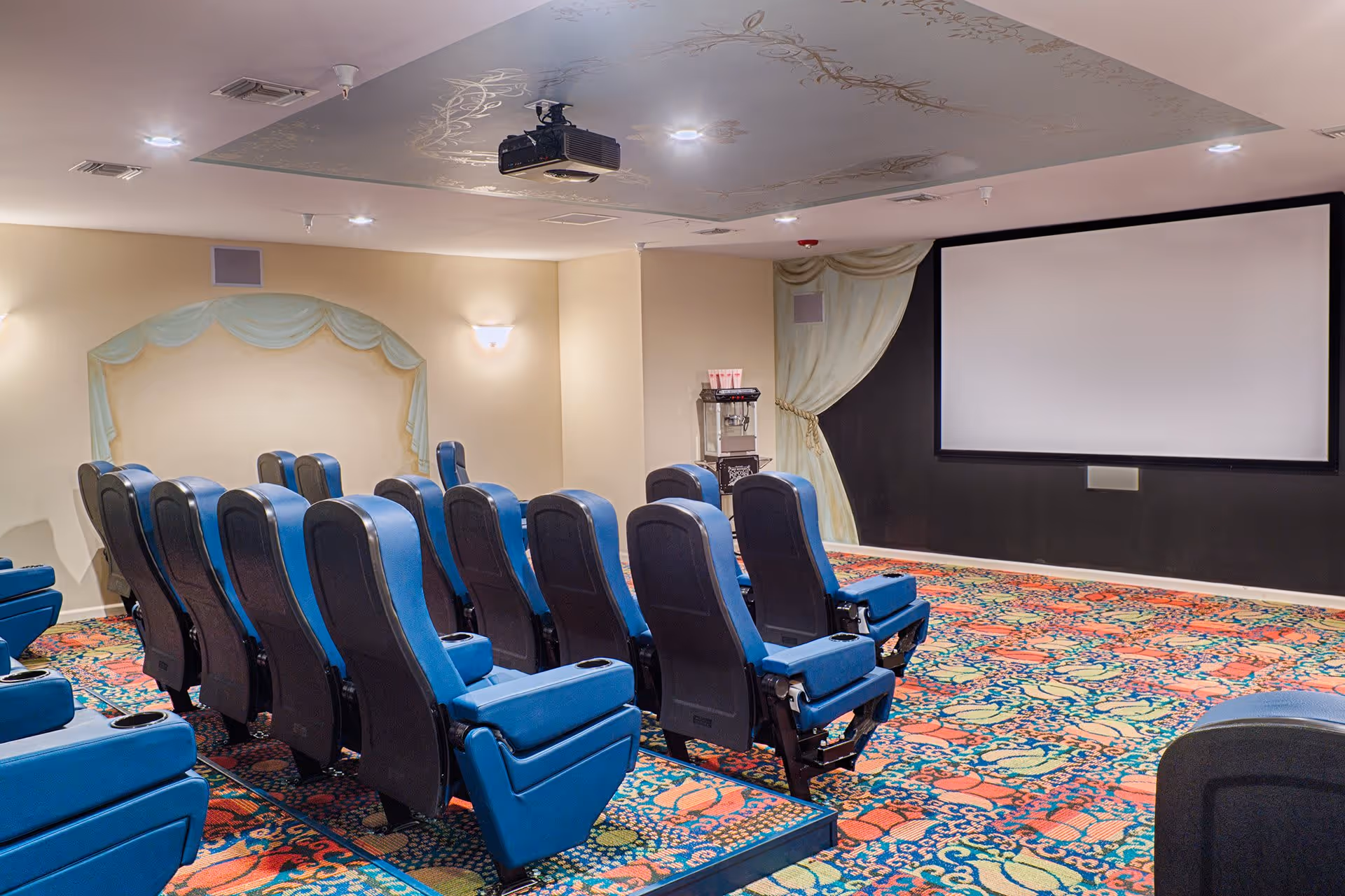 A small theater room with two rows of blue cushioned seats facing a large blank projection screen. The room has patterned carpet with colorful designs, soft wall lighting, and a ceiling-mounted projector. There is a popcorn machine near the screen and decorative curtains on the walls.