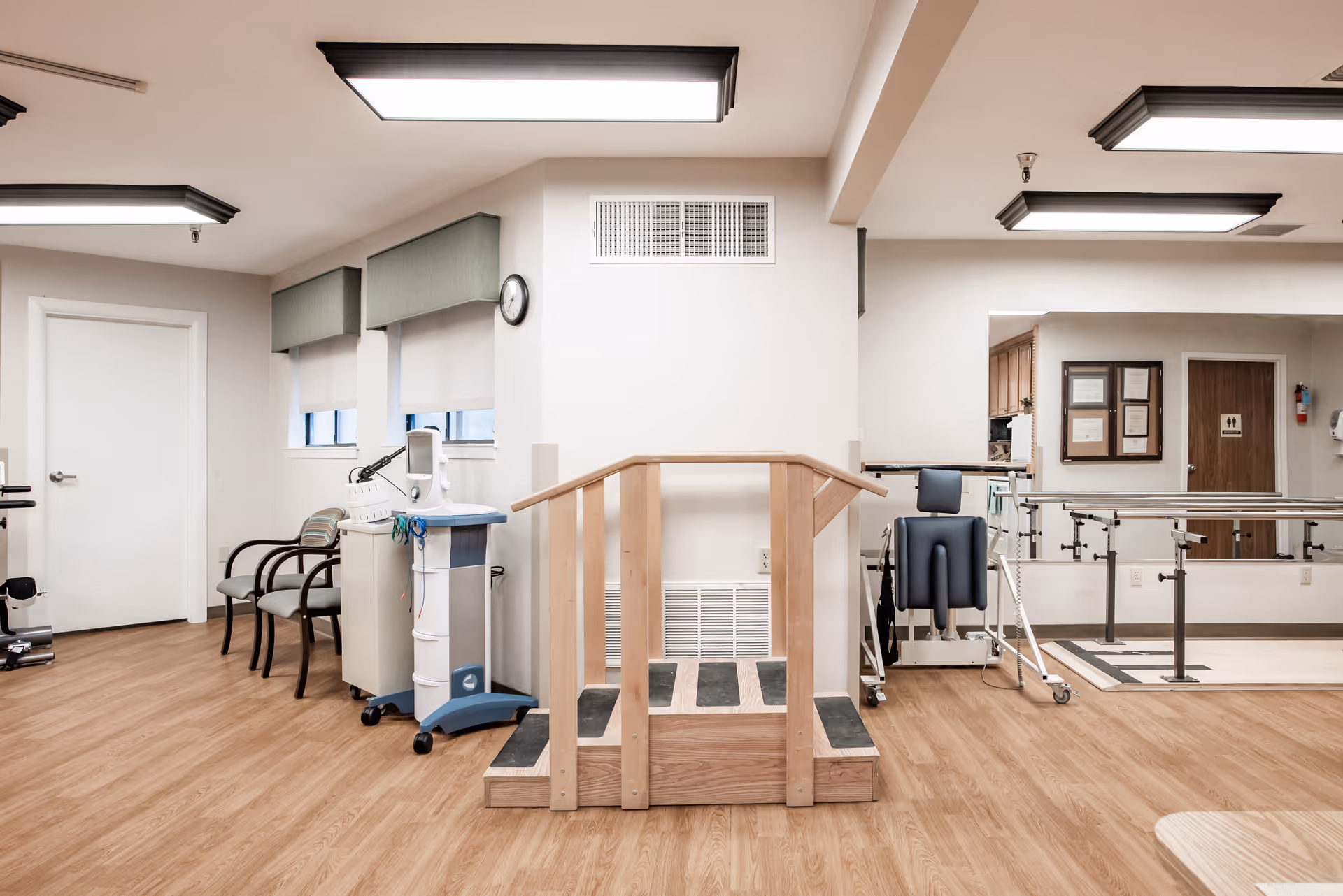 A rehabilitation room with wooden flooring, a small wooden ramp with handrails, parallel bars for walking exercises, chairs, medical equipment, and a clock on the wall. The room is well-lit with ceiling lights and has windows with green valances.