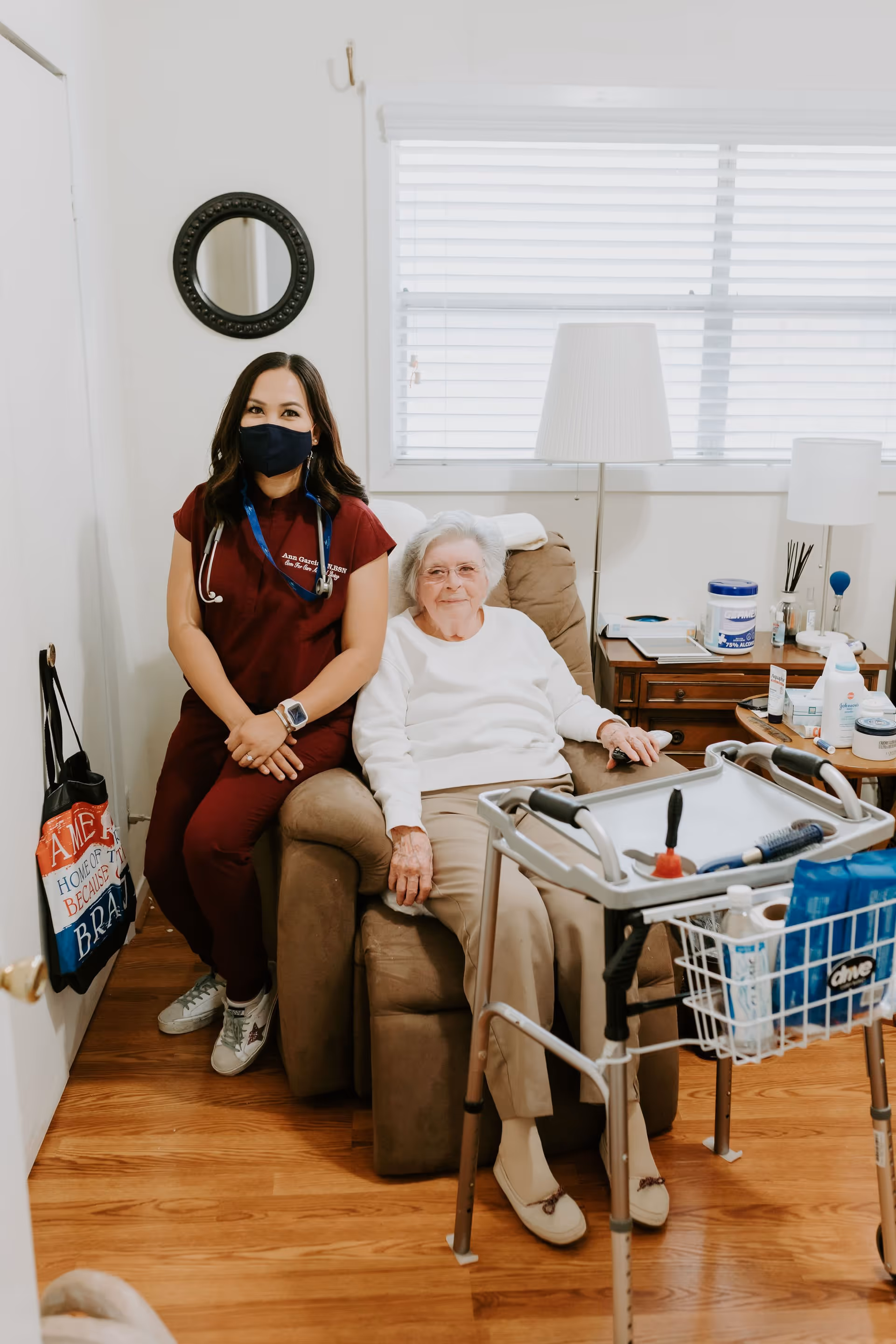 A healthcare worker wearing a mask and maroon scrubs sits next to an elderly woman in a white sweater seated in a recliner chair inside a cozy room. There is a walker with a basket in front of them, a side table with various items, a floor lamp, and a window with blinds in the background.