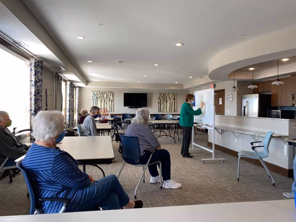 People seated at tables in a community room watching an instructor write on a whiteboard near a kitchenette.