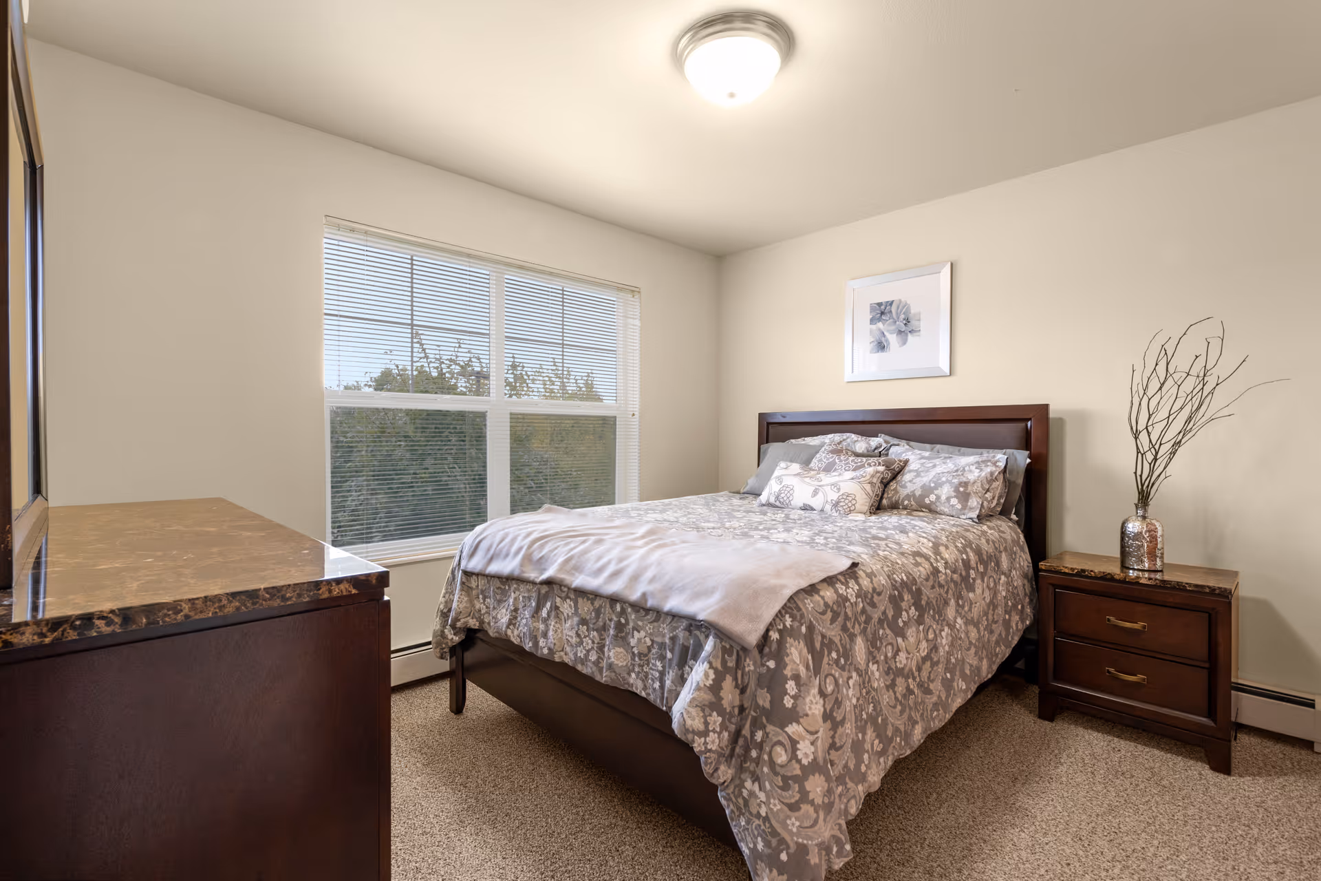 A cozy bedroom with a large window letting in natural light. The room features a bed with a patterned gray and white comforter and multiple pillows. There is a wooden nightstand with two drawers and a decorative vase with branches on it. A matching wooden dresser with a marble-like top is positioned opposite the bed. The walls are painted light beige, and a framed floral artwork hangs above the bed.