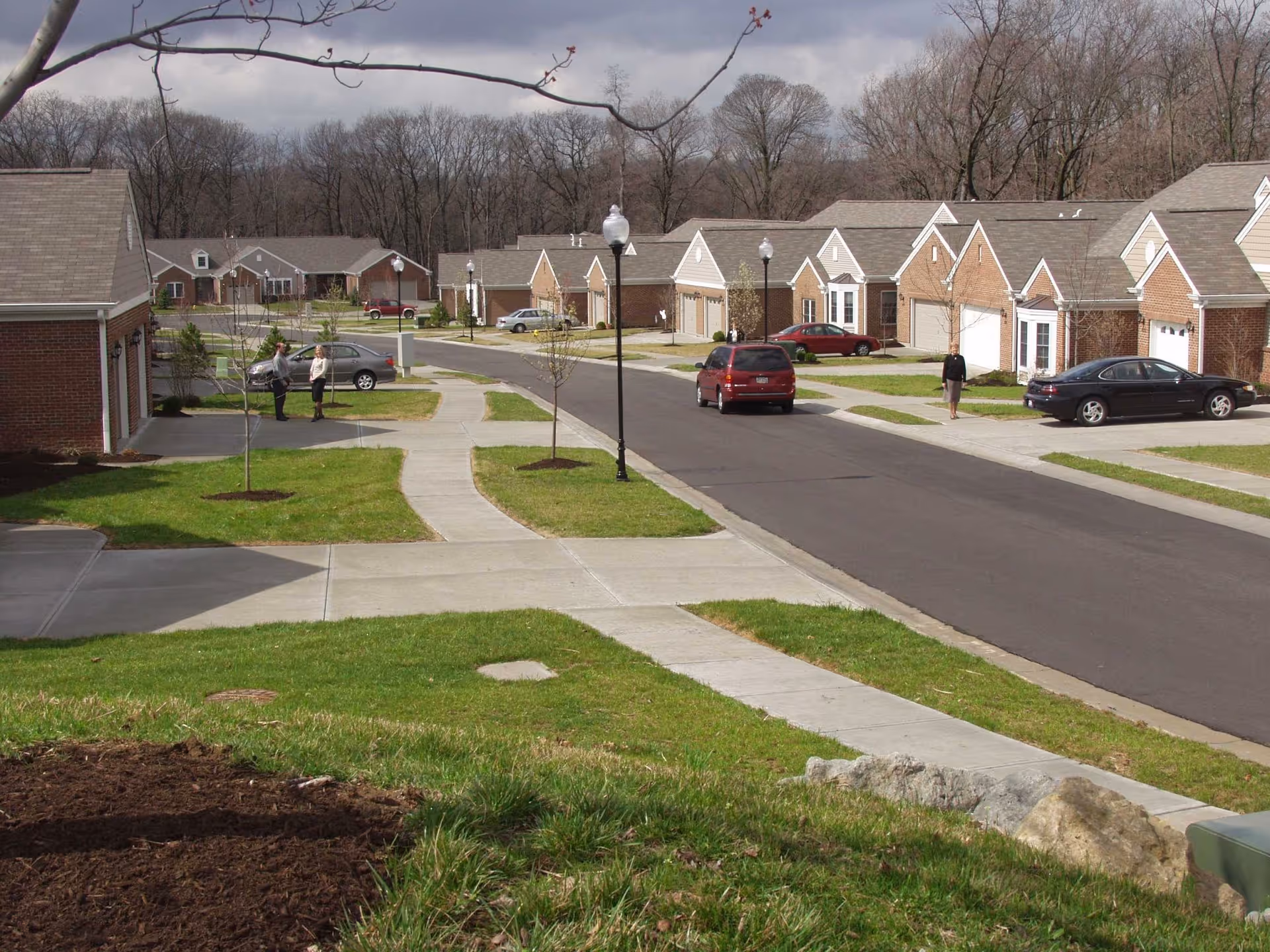 View of a residential community street with single-story brick houses, sidewalks, street lamps, a few parked cars, and three people standing and walking outside. Trees without leaves are visible in the background under a cloudy sky.