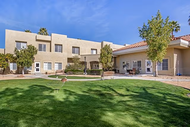 Well-maintained green courtyard with benches in front of a two-story beige senior living building under a blue sky.