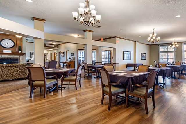 A spacious dining area with multiple tables covered in dark tablecloths and surrounded by wooden chairs with cushioned seats. The room features wooden flooring, beige walls, and several chandeliers hanging from the ceiling. Large windows allow natural light to brighten the space, and a cozy seating area with a fireplace is visible in the background.