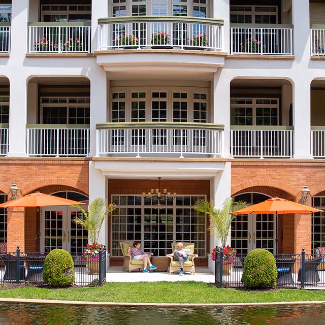 Two elderly people sitting on cushioned chairs under a covered patio area of a multi-story building with balconies. The patio is decorated with potted plants and orange umbrellas, and there is a small body of water in the foreground.