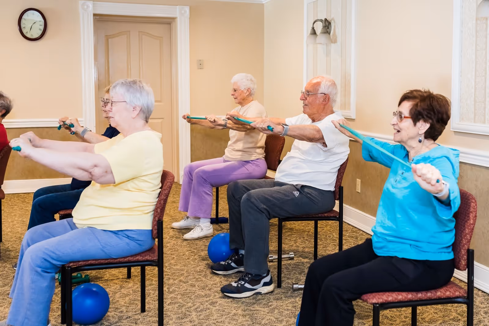 A group of elderly individuals seated on chairs in a room, participating in a seated exercise class using resistance bands. They are extending their arms forward while holding the bands, engaging in a group fitness activity.