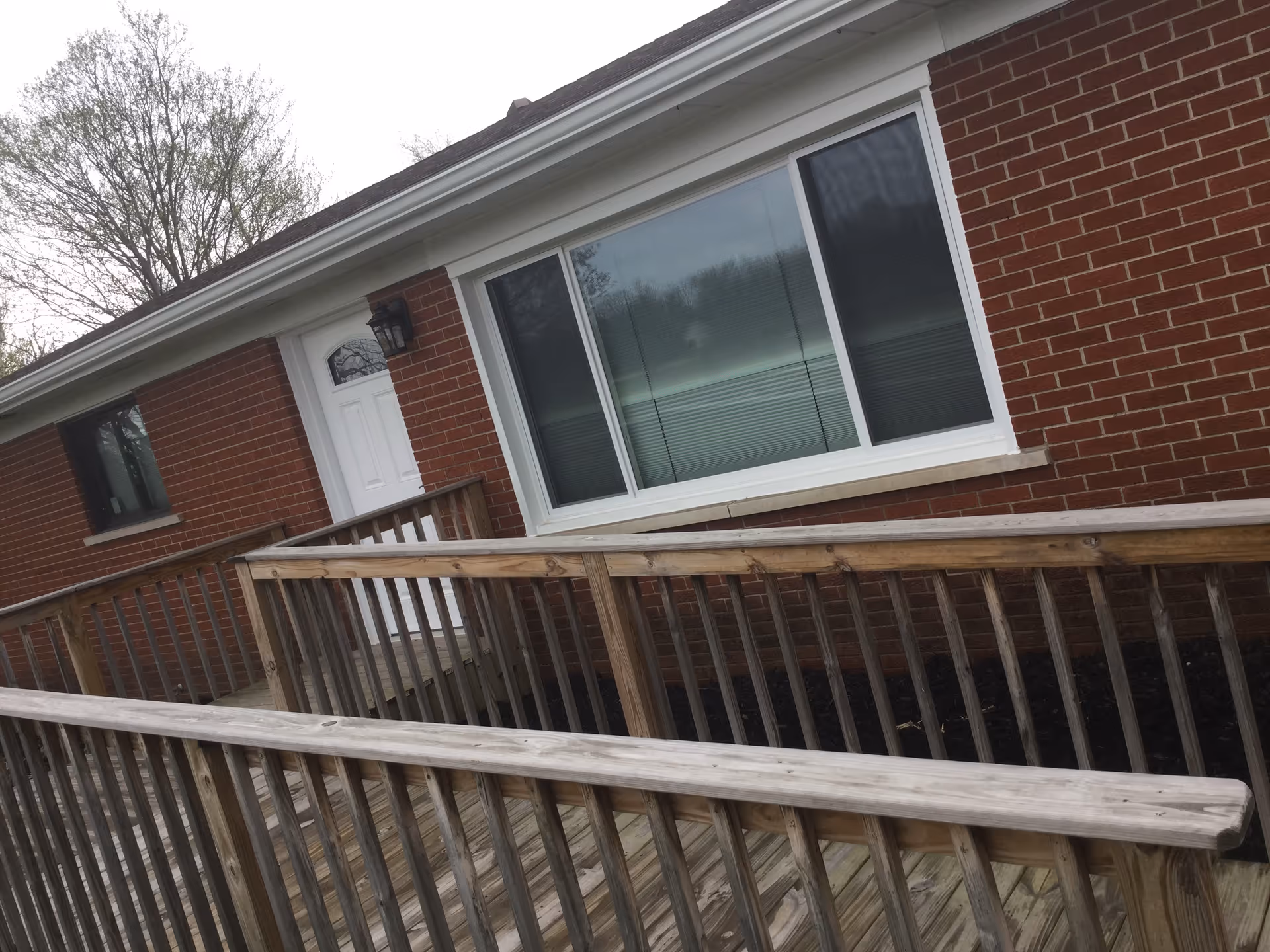 Exterior view of a single-story brick building with a white door and large window. A wooden ramp with railings leads up to the entrance, and trees are visible in the background.