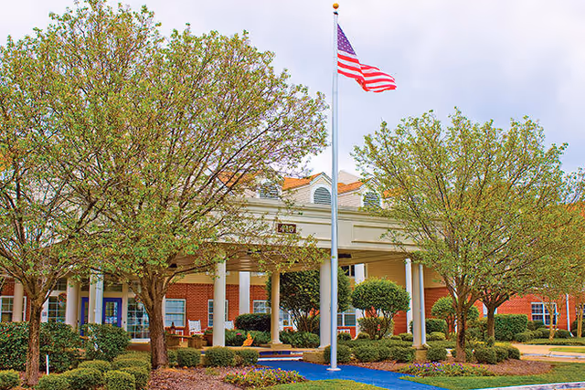 Front exterior view of Ridgeland Place facility with a covered entrance, American flag on a flagpole, and landscaped greenery including trees and bushes.