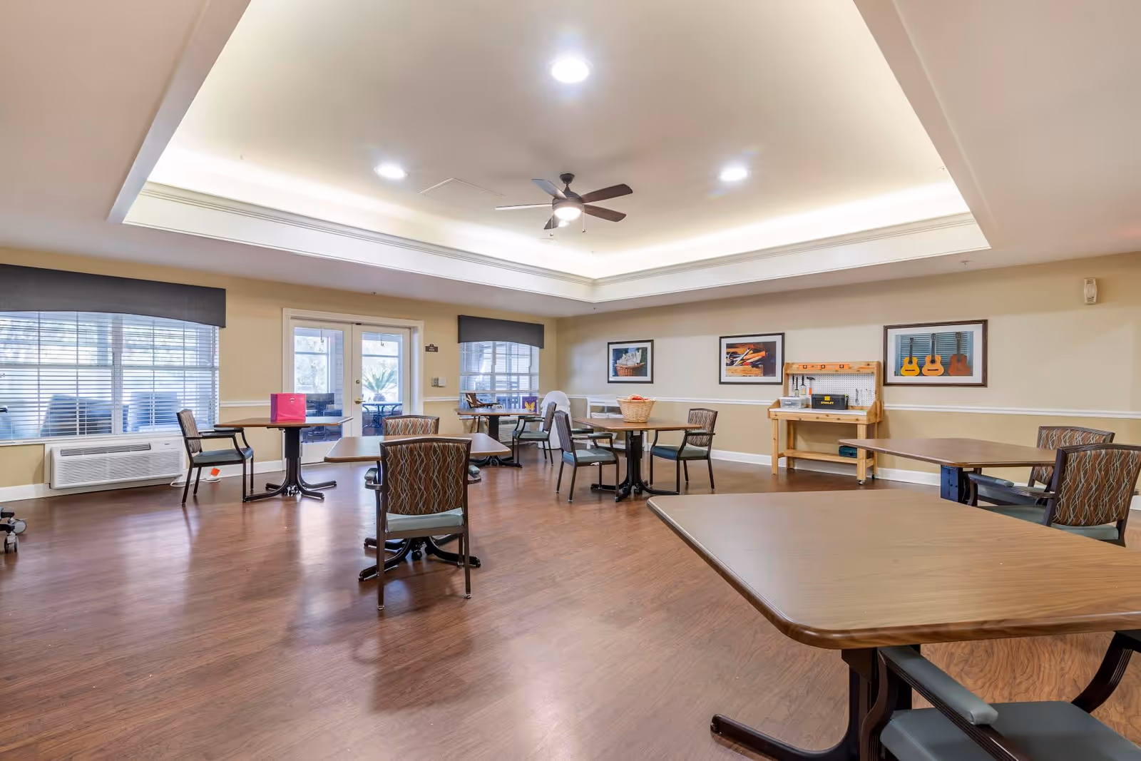 Spacious, well-lit communal activity/dining room with multiple tables and chairs, wood flooring, a ceiling fan, and framed artwork on the walls.