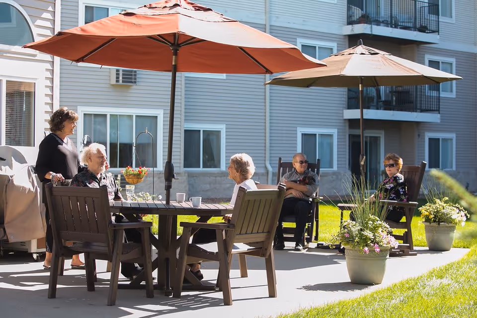 A group of elderly people sitting and standing around outdoor tables with large umbrellas in a sunny patio area of a residential building. There are potted plants with flowers around the seating area, and the building has multiple windows and balconies.