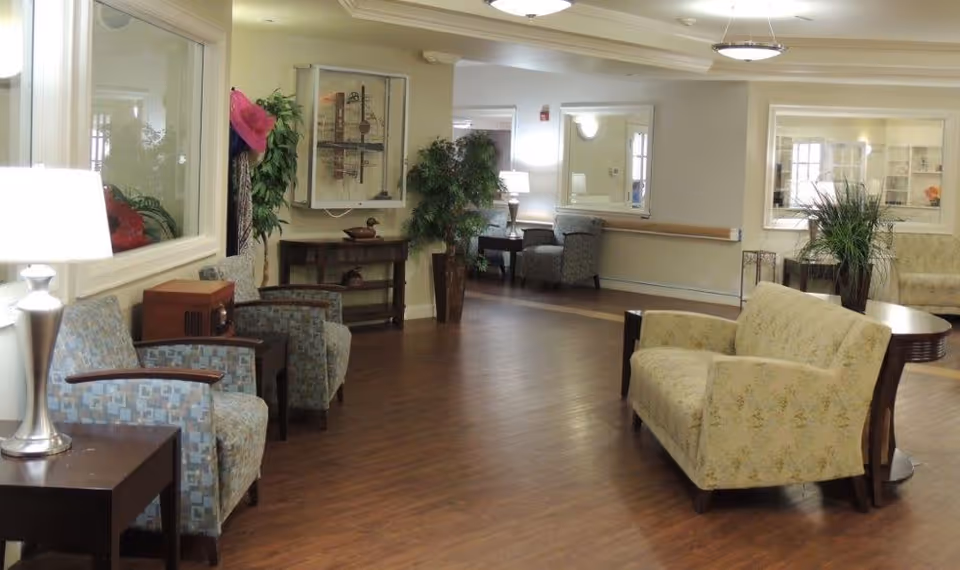 A spacious and well-lit common area in a senior living facility with wooden flooring, multiple armchairs and sofas arranged along the walls, side tables with lamps, potted plants, and framed artwork on the walls.