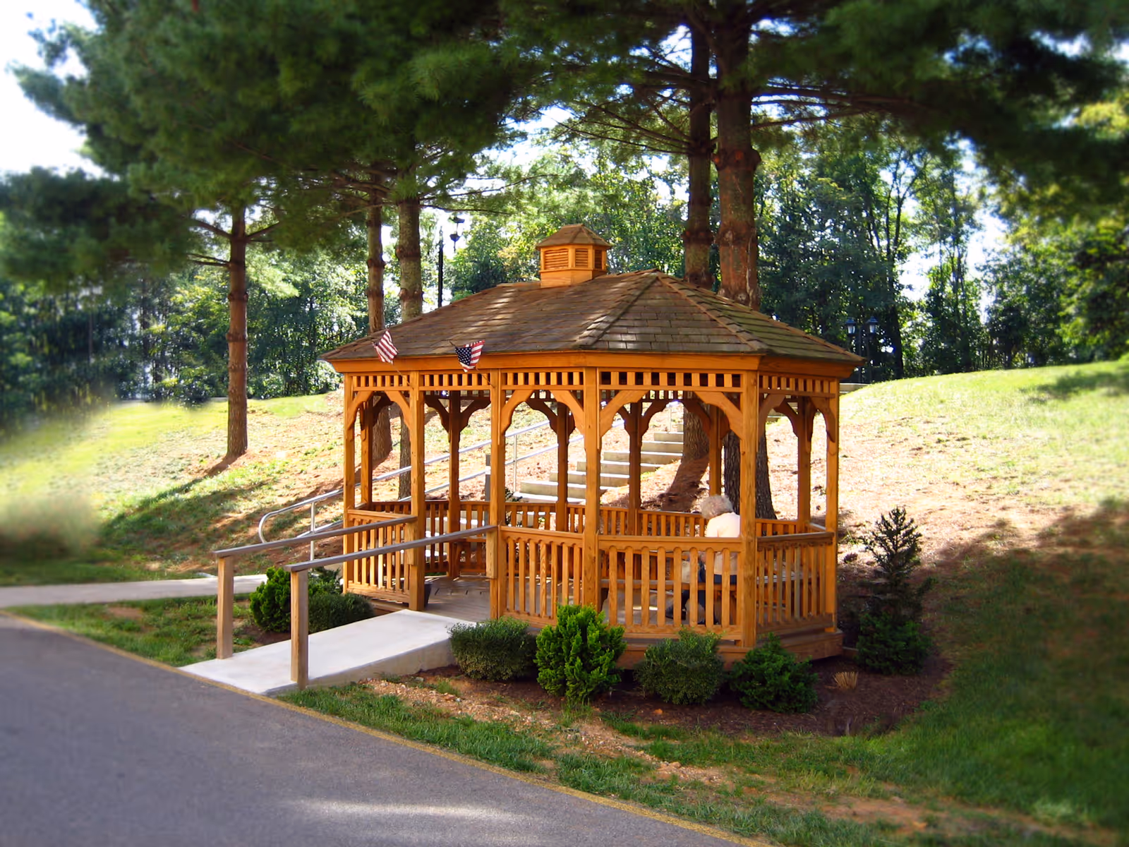 Wooden gazebo with a ramp and benches on a grassy hillside surrounded by trees.