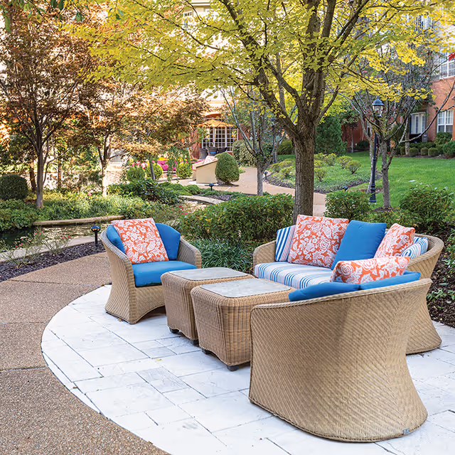 Wicker outdoor seating with blue and orange cushions arranged on a circular stone patio in a landscaped courtyard with trees and walkways.