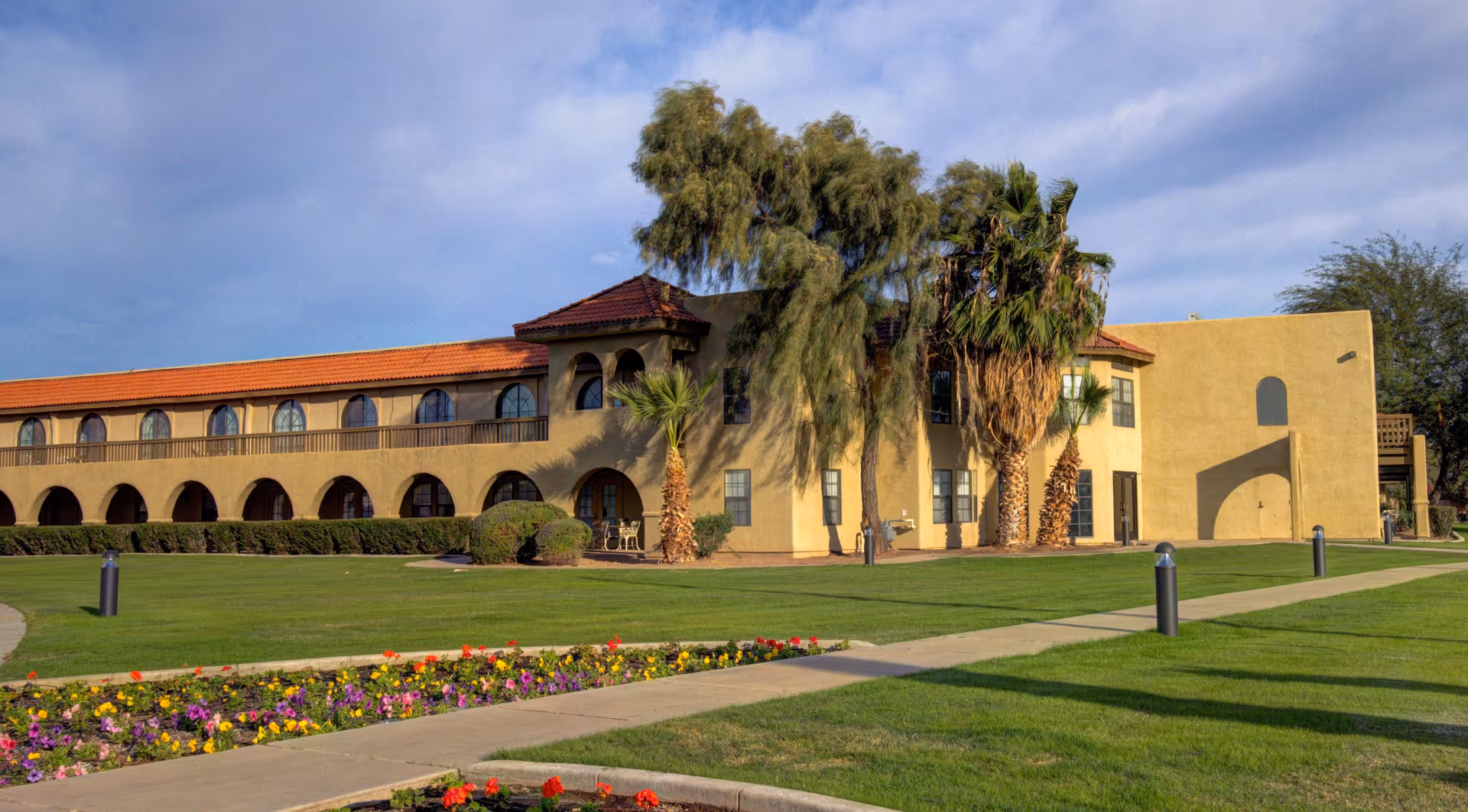 Exterior view of a two-story senior living facility building with a beige stucco finish and red tile roof. The building features arched windows and doorways, surrounded by well-maintained green lawns, palm trees, and colorful flower beds under a partly cloudy sky.
