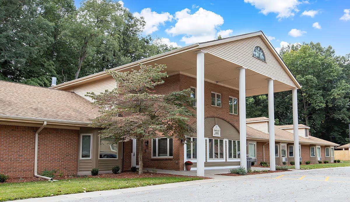 Exterior view of a single-story brick building with a large covered entrance supported by four white columns, surrounded by green trees and a clear blue sky with clouds.