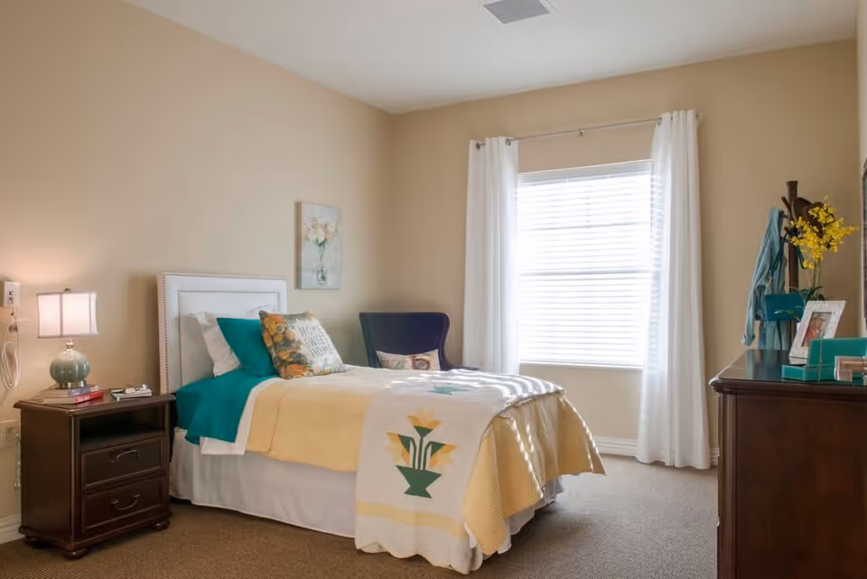 A cozy bedroom with a single bed made with yellow and teal bedding, a white headboard, and decorative pillows including one that says 'Home Sweet Home'. There is a wooden nightstand with a lamp and books on the left side of the bed. A window with white curtains allows natural light to fill the room. On the right side, there is a dark wooden dresser with a vase of yellow flowers, a picture frame, and a coat rack with a teal jacket hanging on it. The walls are painted beige and the floor is carpeted.