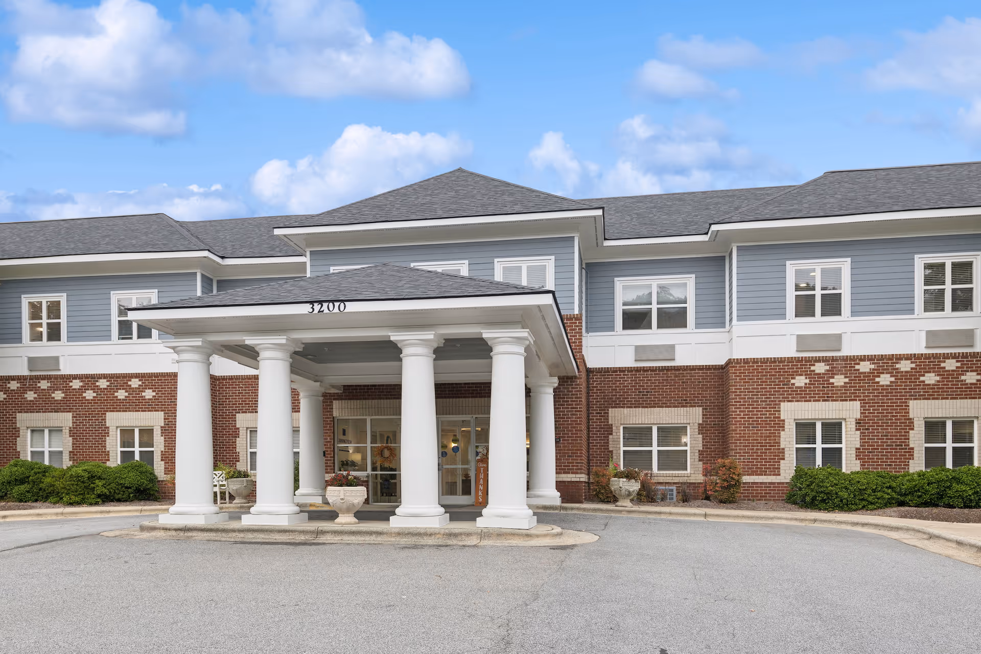 Front exterior view of a senior living facility building with a covered entrance supported by white columns, brick and blue siding walls, multiple windows, and a driveway in front under a partly cloudy sky.
