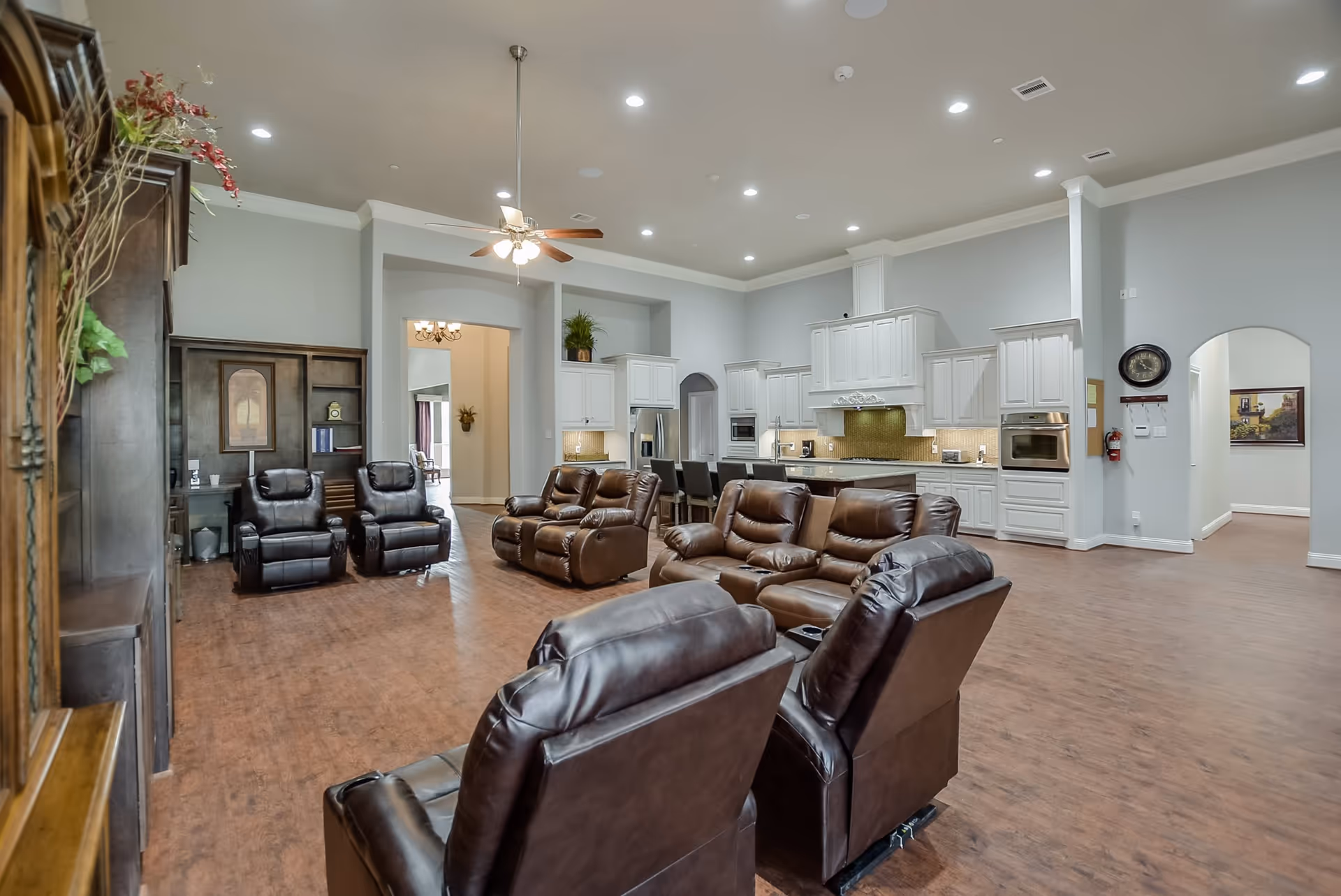Spacious open common area with multiple brown leather recliners facing a white‑cabinet kitchen under high ceilings.