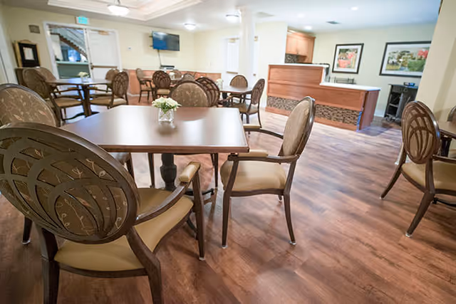 A spacious dining area with multiple wooden tables and cushioned chairs arranged neatly on a wooden floor. The room has light-colored walls, ceiling lights, a small reception desk, and framed artwork on the walls.
