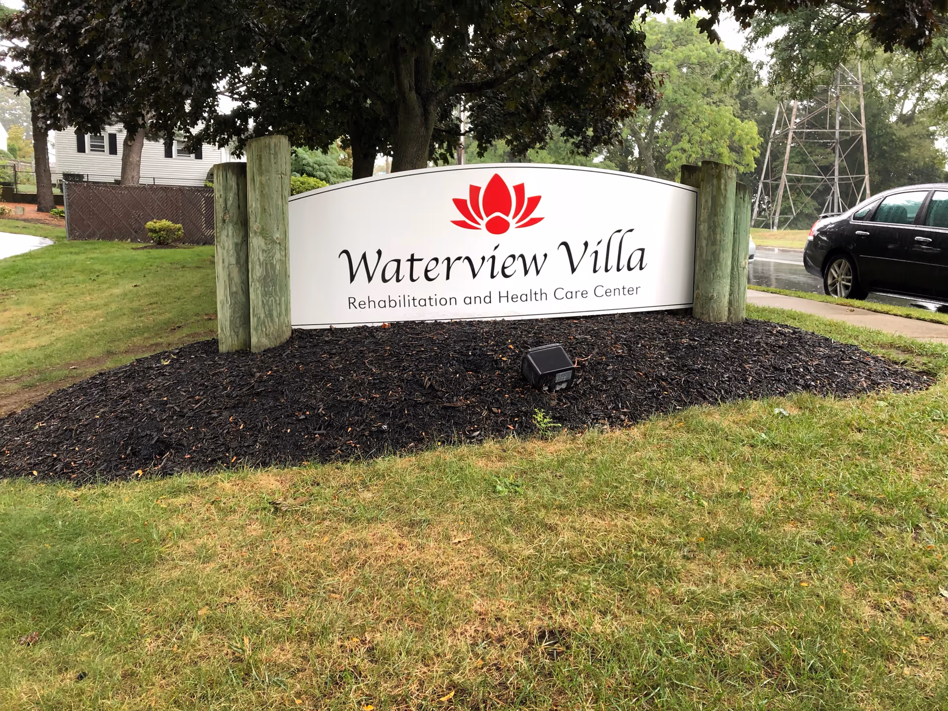 Outdoor sign for Waterview Villa Rehabilitation and Health Care Center surrounded by grass, mulch, and trees, with a black car and a metal tower visible in the background.